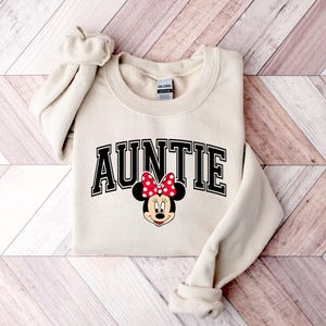 May include: Cream-colored sweatshirt with the word "AUNTIE" in black block letters. A Minnie Mouse graphic with a red polka dot bow is below the text. The sweatshirt has a crew neck and long sleeves.