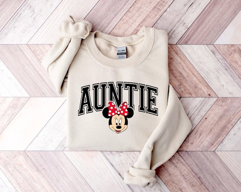 May include: Cream-colored sweatshirt with the word "AUNTIE" in black block letters. A Minnie Mouse graphic with a red polka dot bow is below the text. The sweatshirt has a crew neck and long sleeves.