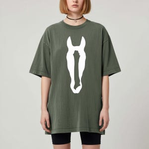 K&ouml;nnte beinhalten: Ein olivgr&uuml;nes Kurzarm-T-Shirt mit einer gro&szlig;en, mittig platzierten wei&szlig;en Grafik, die die Silhouette eines Pferdekopfes zeigt. Das Oberteil hat einen Rundhalsausschnitt und einen lockeren, weit geschnittenen Schnitt in einfarbiger Optik.