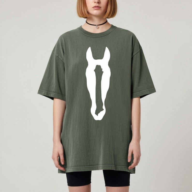 K&ouml;nnte beinhalten: Ein olivgr&uuml;nes Kurzarm-T-Shirt mit einer gro&szlig;en, mittig platzierten wei&szlig;en Grafik, die die Silhouette eines Pferdekopfes zeigt. Das Oberteil hat einen Rundhalsausschnitt und einen lockeren, weit geschnittenen Schnitt in einfarbiger Optik.