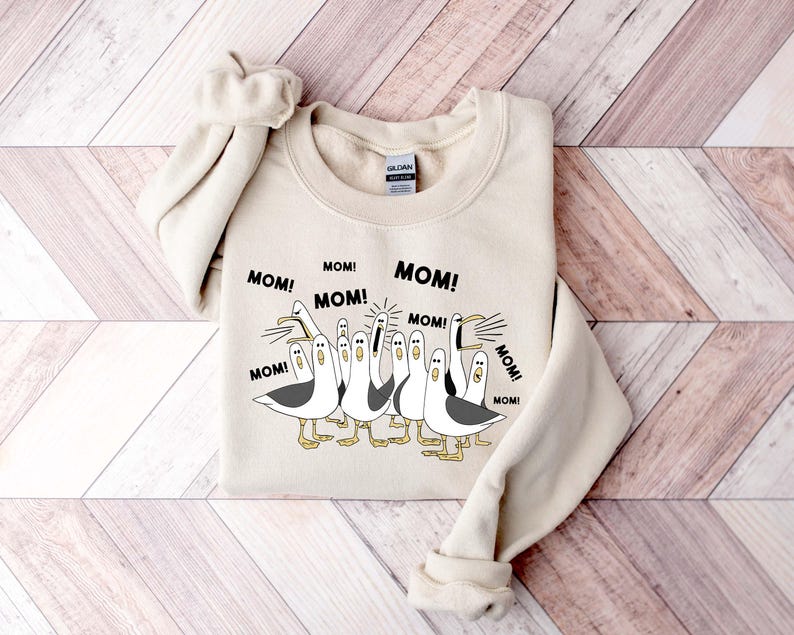 May include: A beige sweatshirt featuring a cartoon design of seagulls with the word "MOM!" repeated around them. The seagulls are in black, white, and gray, with yellow feet. The sweatshirt is laid on a wooden surface.