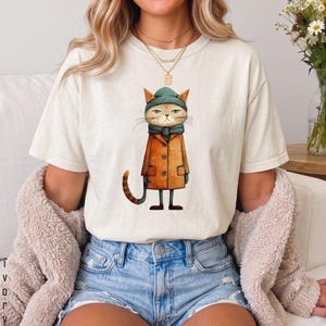 以下が含まれることがあります： オフホワイトの半袖Tシャツ。ティールカラーのビーニーとマフラー、オレンジ色のトレンチコートを身に着けた猫のグラフィックイラストが描かれています。猫は縞模様の尻尾を持ち、無表情です。ブルーのデニムショートパンツと合わせています。