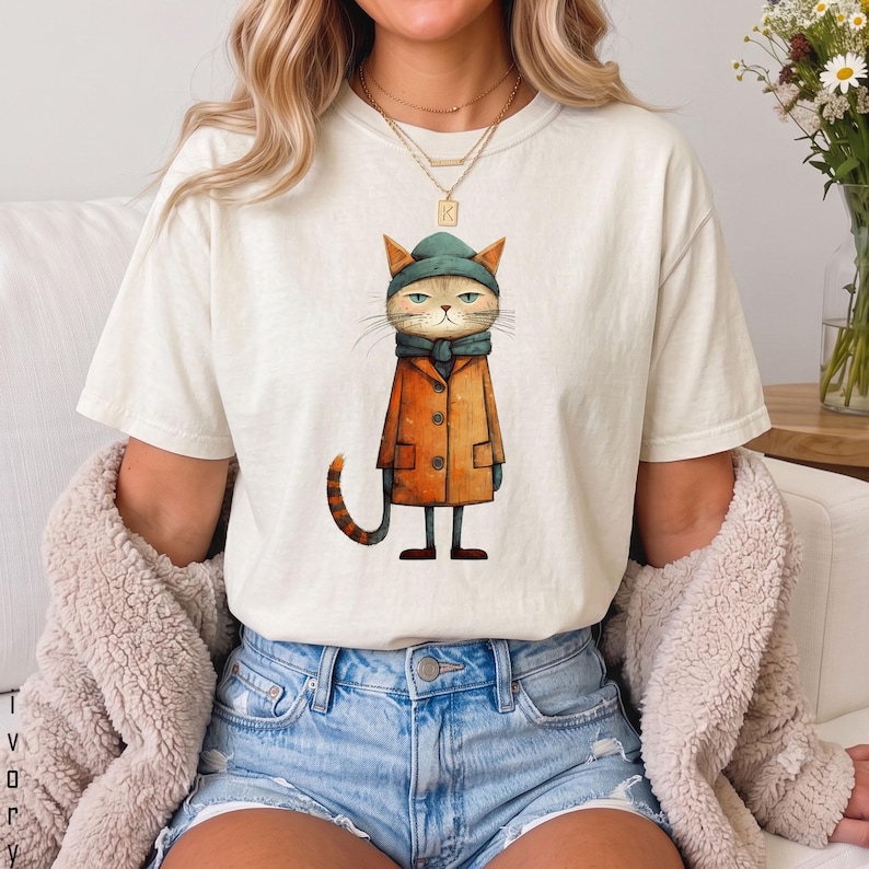 以下が含まれることがあります： オフホワイトの半袖Tシャツ。ティールカラーのビーニーとマフラー、オレンジ色のトレンチコートを身に着けた猫のグラフィックイラストが描かれています。猫は縞模様の尻尾を持ち、無表情です。ブルーのデニムショートパンツと合わせています。
