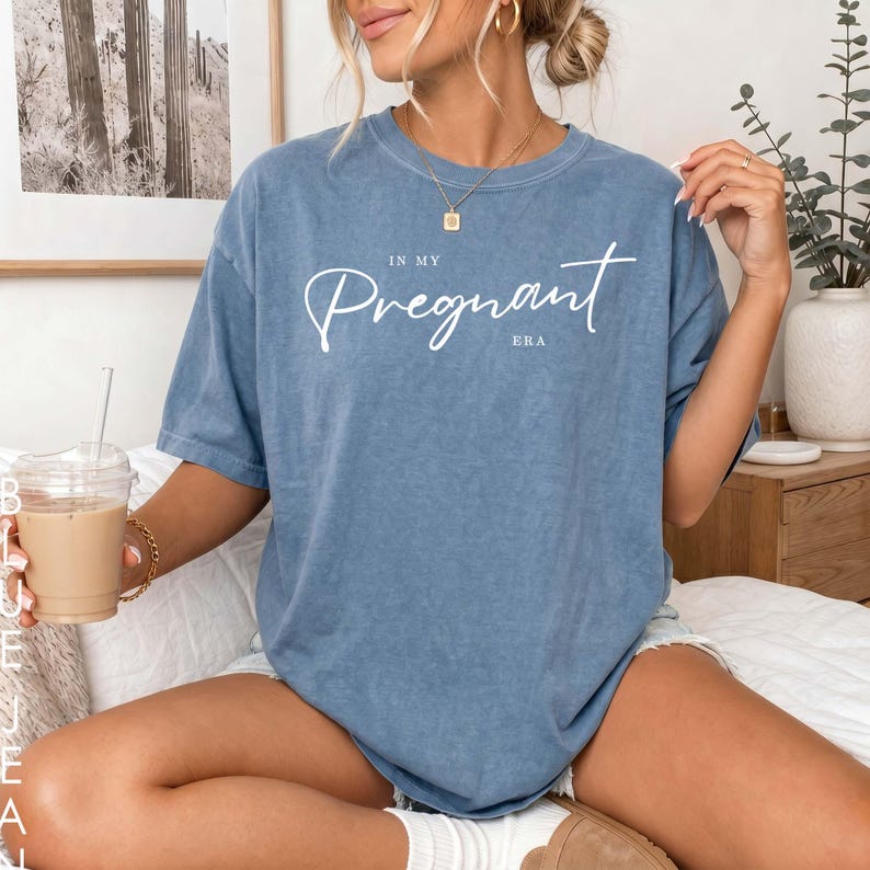 May include: A blue short-sleeved t-shirt with a washed-out finish featuring the white text "IN MY PREGNANT ERA" printed across the chest in a combination of a cursive script font and a simple sans-serif typeface for the word "ERA".