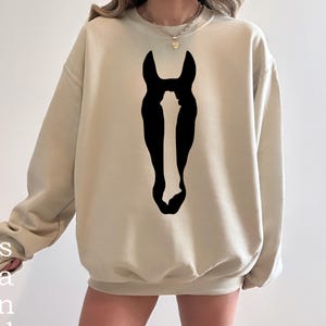K&ouml;nnte beinhalten: Ein beigefarbenes Sweatshirt mit Rundhalsausschnitt und einer gro&szlig;en, mittig platzierten schwarzen Grafik einer Pferdekopf-Silhouette. Der lang&auml;rmelige Pullover verf&uuml;gt &uuml;ber gerippte B&uuml;ndchen an Kragen, &Auml;rmeln und Saum f&uuml;r eine lockere Passform.