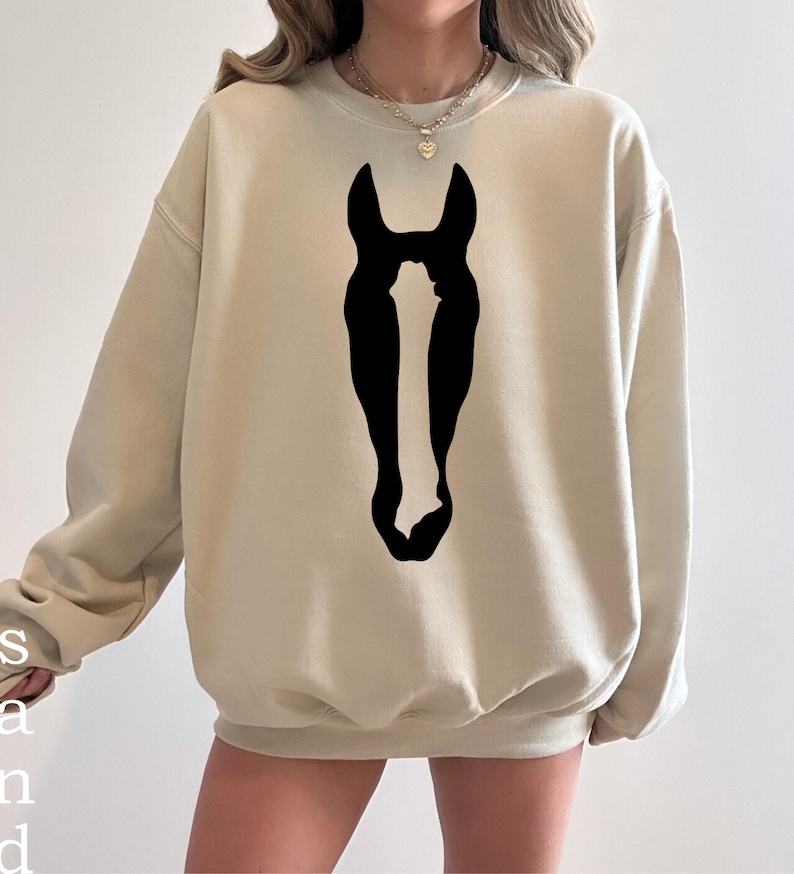 K&ouml;nnte beinhalten: Ein beigefarbenes Sweatshirt mit Rundhalsausschnitt und einer gro&szlig;en, mittig platzierten schwarzen Grafik einer Pferdekopf-Silhouette. Der lang&auml;rmelige Pullover verf&uuml;gt &uuml;ber gerippte B&uuml;ndchen an Kragen, &Auml;rmeln und Saum f&uuml;r eine lockere Passform.
