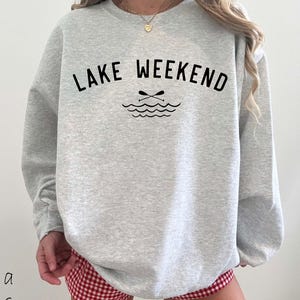 May include: A light gray crewneck sweatshirt featuring the black text "LAKE WEEKEND" arched over a graphic of two crossed oars and stylized water waves. The garment has long sleeves and a ribbed neckline, paired with red and white gingham shorts.