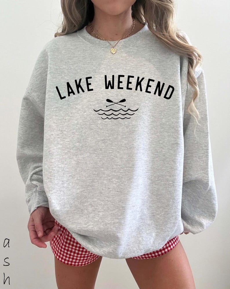 May include: A light gray crewneck sweatshirt featuring the black text "LAKE WEEKEND" arched over a graphic of two crossed oars and stylized water waves. The garment has long sleeves and a ribbed neckline, paired with red and white gingham shorts.