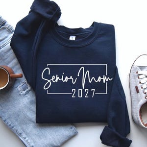 Może przedstawiać: Granatowa bluza z okrągłym dekoltem z białym napisem Senior Mom 2027 w prostokątnej ramce. Bluza jest pokazana obok jasnoniebieskich dżins&oacute;w z przetarciami, brązowego ceramicznego kubka i szarych but&oacute;w sportowych.
