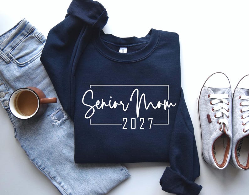Może przedstawiać: Granatowa bluza z okrągłym dekoltem z białym napisem Senior Mom 2027 w prostokątnej ramce. Bluza jest pokazana obok jasnoniebieskich dżins&oacute;w z przetarciami, brązowego ceramicznego kubka i szarych but&oacute;w sportowych.