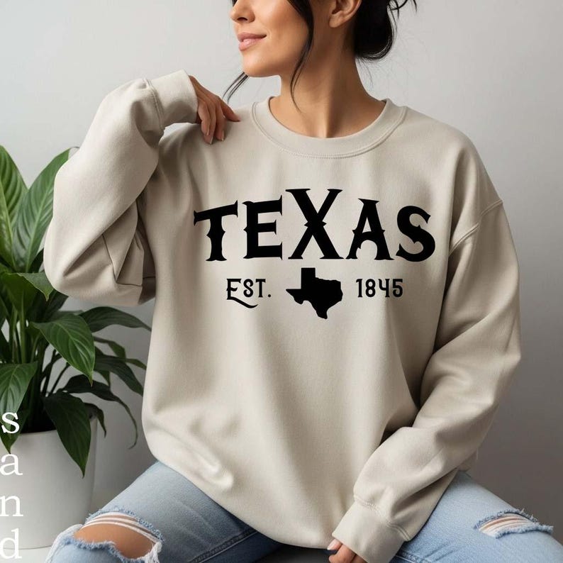 K&ouml;nnte beinhalten: Ein beigefarbenes Sweatshirt mit Rundhalsausschnitt und schwarzem Aufdruck TEXAS EST. 1845 sowie einer schwarzen Silhouette des Bundesstaates Texas zwischen der Abk&uuml;rzung und der Jahreszahl. Das lang&auml;rmelige Kleidungsst&uuml;ck hat gerippte B&uuml;ndchen.
