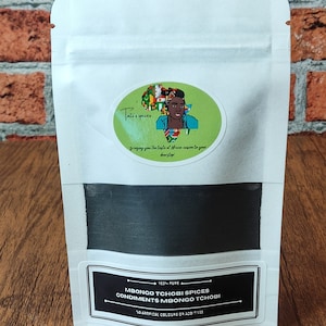 Könnte beinhalten: Ein weißer wiederverschließbarer Beutel mit einem schwarzen Etikett, auf dem "100% PURE MBONGO TCHOBI SPICES CONDIMENTS MBONGO TCHOBI NO ARTIFICAL COLOURS OR ADDITIVES" steht. Der Beutel enthält ein schwarzes Pulver.