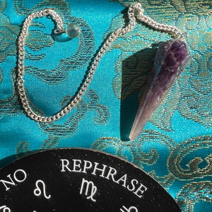 May include: A silver chain pendulum with a faceted amethyst crystal point and a clear crystal bead. The pendulum is displayed on a teal patterned fabric, with a black round board that says "NO REPHRASE" and zodiac symbols.
