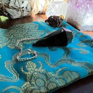 May include: Black obsidian pendulum with a silver chain on a blue and gold patterned fabric pouch.