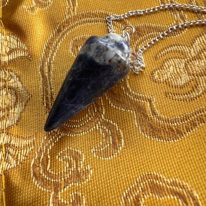 May include: A blue and white crystal pendulum with a silver chain. The pendulum is hanging on a gold fabric background.
