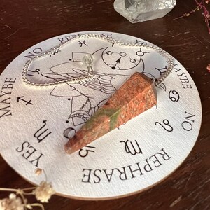 May include: A red and green crystal pendulum on a silver chain hanging over a white circular board with the words "YES", "MAYBE", "NO", "REPHRASE" and astrological symbols.