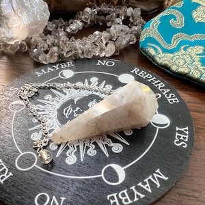 May include: A white crystal pendulum on a black circular board with the words "Rephrase", "Yes", "Maybe", "No", and "Rephrase" printed around the edge. The board is surrounded by clear quartz crystals.
