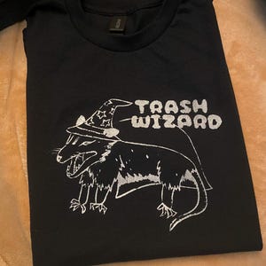 May include: Black t-shirt with a white graphic of a possum wearing a wizard hat. The text "Trash Wizard" is printed above the possum.