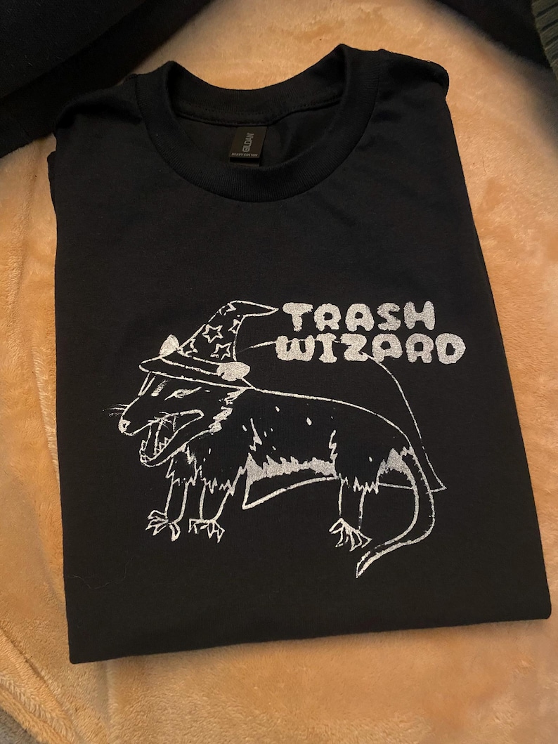 May include: Black t-shirt with a white graphic of a possum wearing a wizard hat. The text "Trash Wizard" is printed above the possum.