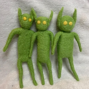 May include: Three needle-felted figures in a vibrant lime green color. Each figure features a rounded head with two pointed ears, a slender torso, two arms, and two legs. The eyes are represented by two small, circular, bright yellow felted dots.