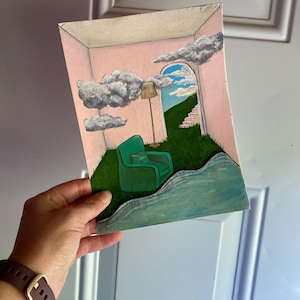 May include: A surreal painting shows a room with a green armchair, a lamp, and a doorway leading to a grassy hill and sky. Fluffy clouds float in the pink-walled room. The painting is held against a white door.