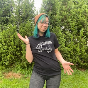 May include: A person with teal hair wearing a brown beanie and a black t-shirt with a white graphic of a lizard wearing a wizard hat and the text "Trash Wizard".