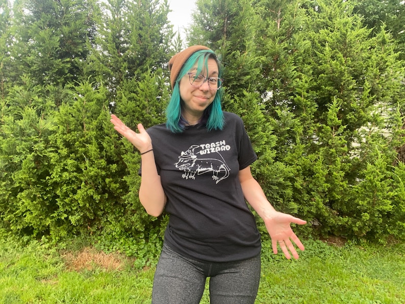 May include: A person with teal hair wearing a brown beanie and a black t-shirt with a white graphic of a lizard wearing a wizard hat and the text "Trash Wizard".