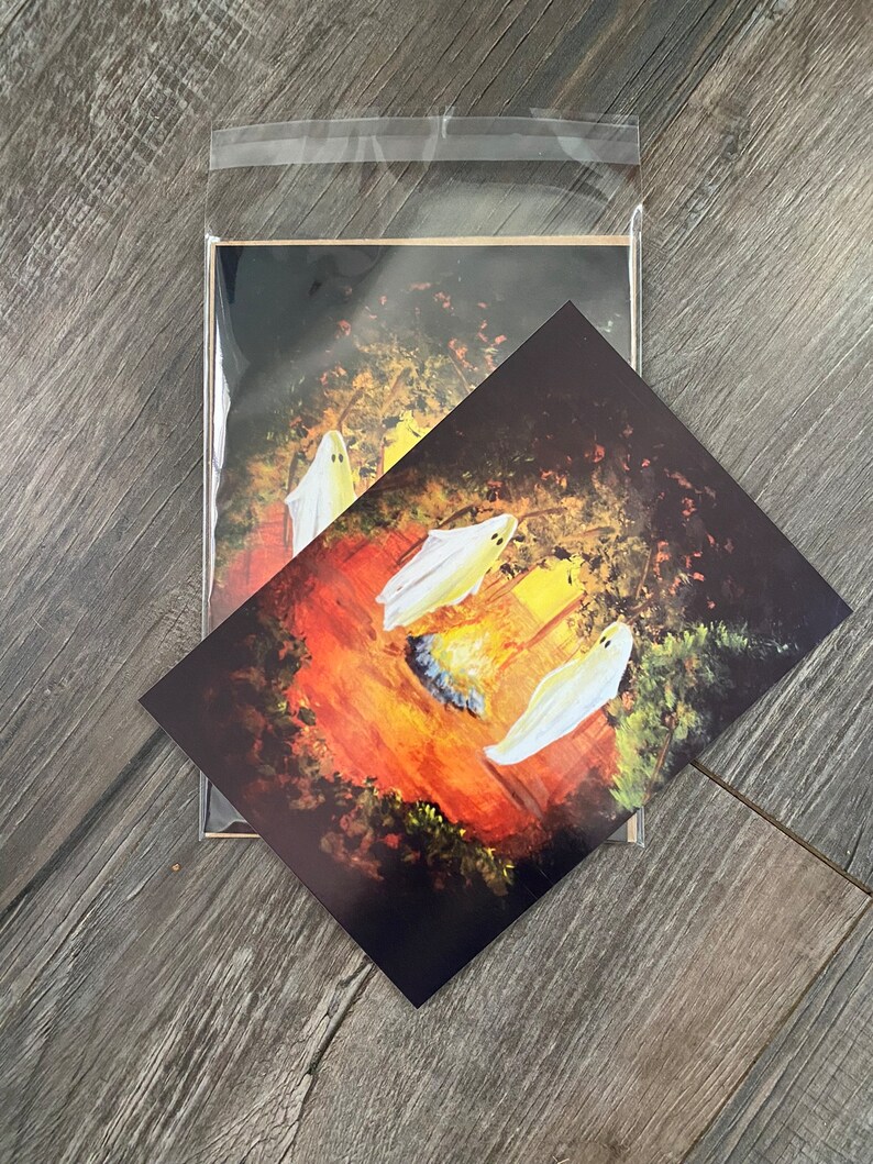 K&ouml;nnte beinhalten: Drei wei&szlig;e Geister versammeln sich um ein Lagerfeuer in einer dunklen Waldkulisse. Die Geister sind in einem verspielten Stil mit leuchtenden Augen und einem warmen orangenen Schein vom Feuer gemalt.