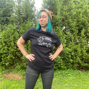 May include: A person wearing a black t-shirt with a white graphic of a toad with a crown and the text "TOAD WIZARD". The person is wearing a brown beanie and has blue hair.