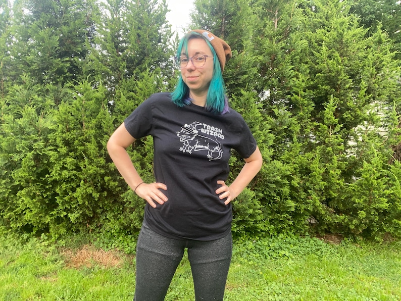 May include: A person wearing a black t-shirt with a white graphic of a toad with a crown and the text "TOAD WIZARD". The person is wearing a brown beanie and has blue hair.