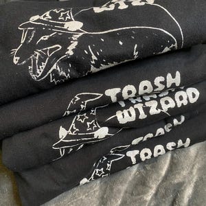 May include: A stack of black t-shirts with white graphic designs. The top shirt features a cartoon creature wearing a hat with the text "Trash Wizard". The other shirts feature the text "Trash Wizard" in a similar style.