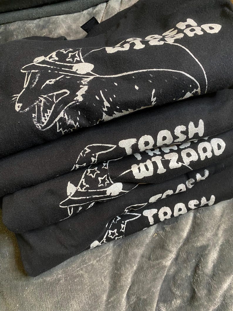 May include: A stack of black t-shirts with white graphic designs. The top shirt features a cartoon creature wearing a hat with the text "Trash Wizard". The other shirts feature the text "Trash Wizard" in a similar style.