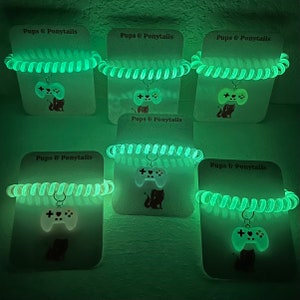 May include: Five glow-in-the-dark green bracelets with a white game controller charm. Each bracelet has a white card with the text "Pups & Ponytails" printed on it.