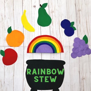 Puede incluir: Un arcoíris de fieltro con un centro morado, una caldera de fieltro negro con el texto "RAINBOW STEW", y fruta de fieltro en varios colores, incluyendo rojo, naranja, amarillo, verde y morado.