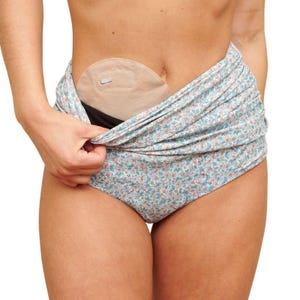 Pode incluir: Roupa interior com estampa floral azul claro com uma bolsa de ostomia bege. A roupa interior é ligeiramente puxada para baixo, revelando a bolsa de ostomia. A bolsa é bege e tem uma pequena abertura.