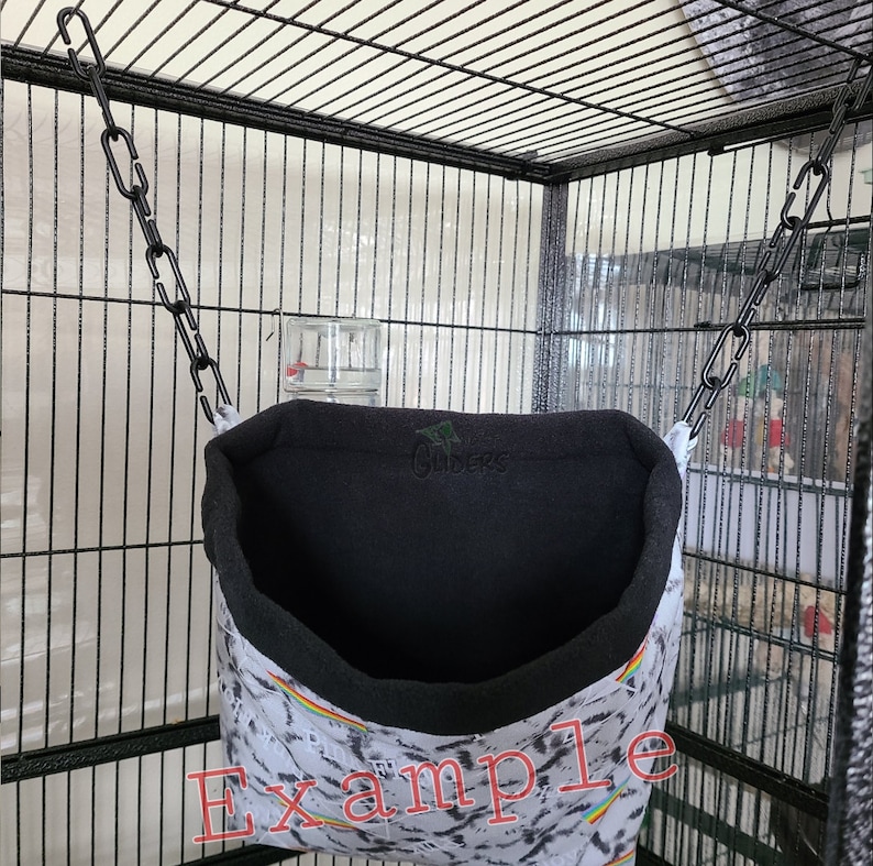 May include: A black and white patterned hanging bird hammock with a black fleece lining. The hammock is attached to a black chain and hangs inside a black metal cage. The text "Example" is written on the hammock.