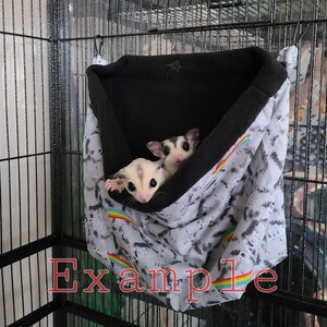 May include: Two sugar gliders are nestled in a gray and white patterned hanging pouch with a black fleece lining. The pouch has a rainbow stripe across the bottom.
