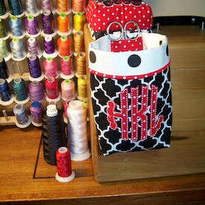 May include: A black and white fabric storage container with red polka dots and a red monogram. The container is filled with spools of thread and a pair of scissors.