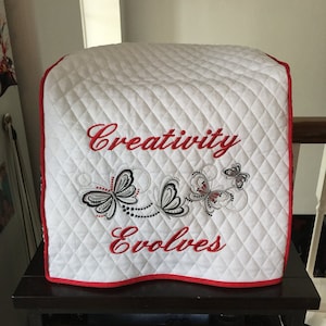 May include: White quilted cover with red trim for a kitchen appliance. The cover features black and grey embroidered butterflies and the text "Creativity Evolves" in red.