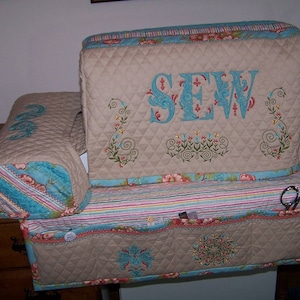 May include: Beige quilted sewing machine cover with a turquoise trim and the word "SEW" embroidered in turquoise. The cover has a matching turquoise and beige striped pocket for storing sewing supplies.