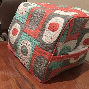 May include: A quilted fabric cover for a bread machine. The cover is made of patchwork fabric in shades of gray, teal, and coral. The fabric has a variety of patterns and includes text that reads "Say My Name" and "Thank You For The Good Times".