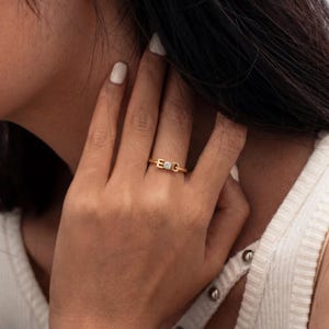 Puede incluir: Un anillo dorado con las letras "E" y "G" y una pequeña gema cuadrada. El anillo está en un dedo. La persona lleva una camiseta blanca. El anillo es una joya.
