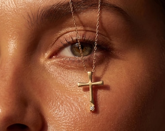 May include: A gold-toned metal cross pendant necklace featuring a delicate chain and a single small, clear, round-cut gemstone set at the base of the cross. The pendant hangs vertically, showcasing a polished, smooth surface and a simple design.
