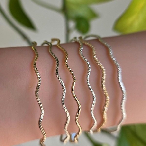 Puede incluir: Una colección de brazaletes ondulados dorados y plateados. Las pulseras tienen un aspecto texturizado y martillado, con un diseño delicado y fluido. Las joyas se muestran en un brazo, con follaje verde en el fondo.