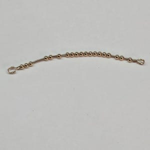 May include: A silver-coloured beaded chain with small, round beads. The chain has a hook on each end. The beads are evenly spaced along the chain, which is slightly curved. The chain is likely used for a light fixture or similar item.
