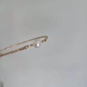 May include: Delicate gold-toned bracelet featuring a single, luminous pearl. The bracelet has a textured band and a simple, elegant design. This piece of jewelry is perfect for adding a touch of sophistication to any outfit.