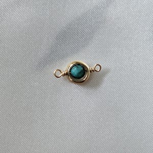 May include: A small, gold-colored jewelry component with a turquoise-colored stone in the center. The component has a circular design around the stone and loops on either end. The background is a white, textured fabric.