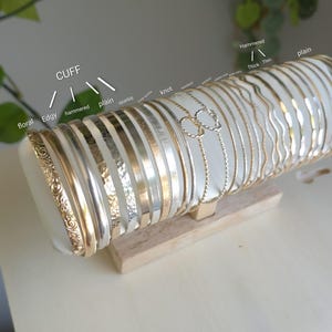 May include: A collection of metal bangles and cuffs displayed on a cylindrical holder. The bracelets feature various textures including floral, hammered, plain, sparkle, and knot designs, with labels such as CUFF, floral, Edgy, hammered, plain, sparkle, knot, and wavy.