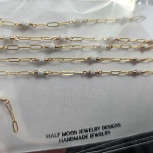 Puede incluir: Cinco cadenas de oro con peque&ntilde;as cuentas blancas, cada cadena tiene una longitud diferente. Las cadenas est&aacute;n sobre un fondo blanco. El texto "HALF MOON JEWELRY DESIGNS HANDMADE JEWELRY" est&aacute; en la parte inferior de la imagen.
