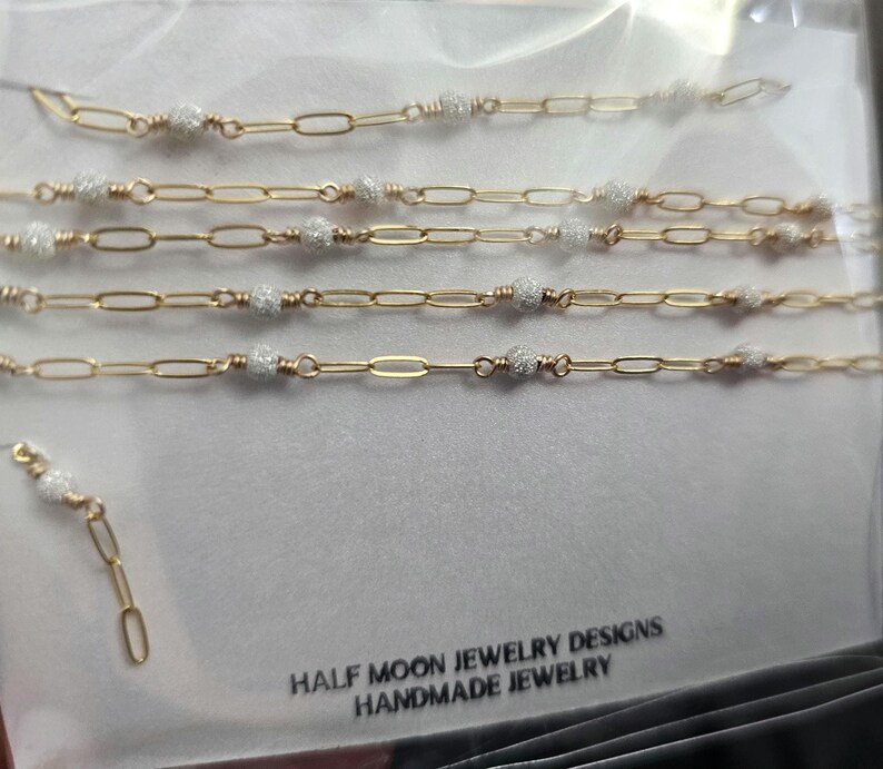 Puede incluir: Cinco cadenas de oro con peque&ntilde;as cuentas blancas, cada cadena tiene una longitud diferente. Las cadenas est&aacute;n sobre un fondo blanco. El texto "HALF MOON JEWELRY DESIGNS HANDMADE JEWELRY" est&aacute; en la parte inferior de la imagen.